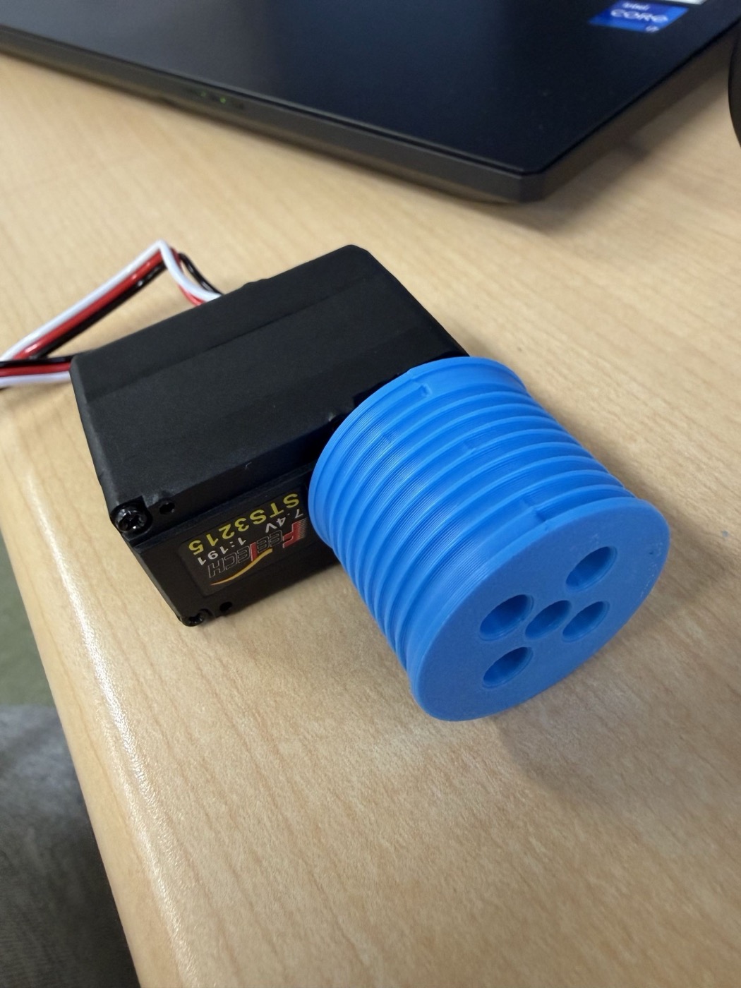 机の上に置かれた黒いFeetech STS3215サーボに、青色の3Dプリント製ドラムが取り付けられている。ドラムの側面には螺旋状の溝が彫られ、端面には複数の穴が並んでいる