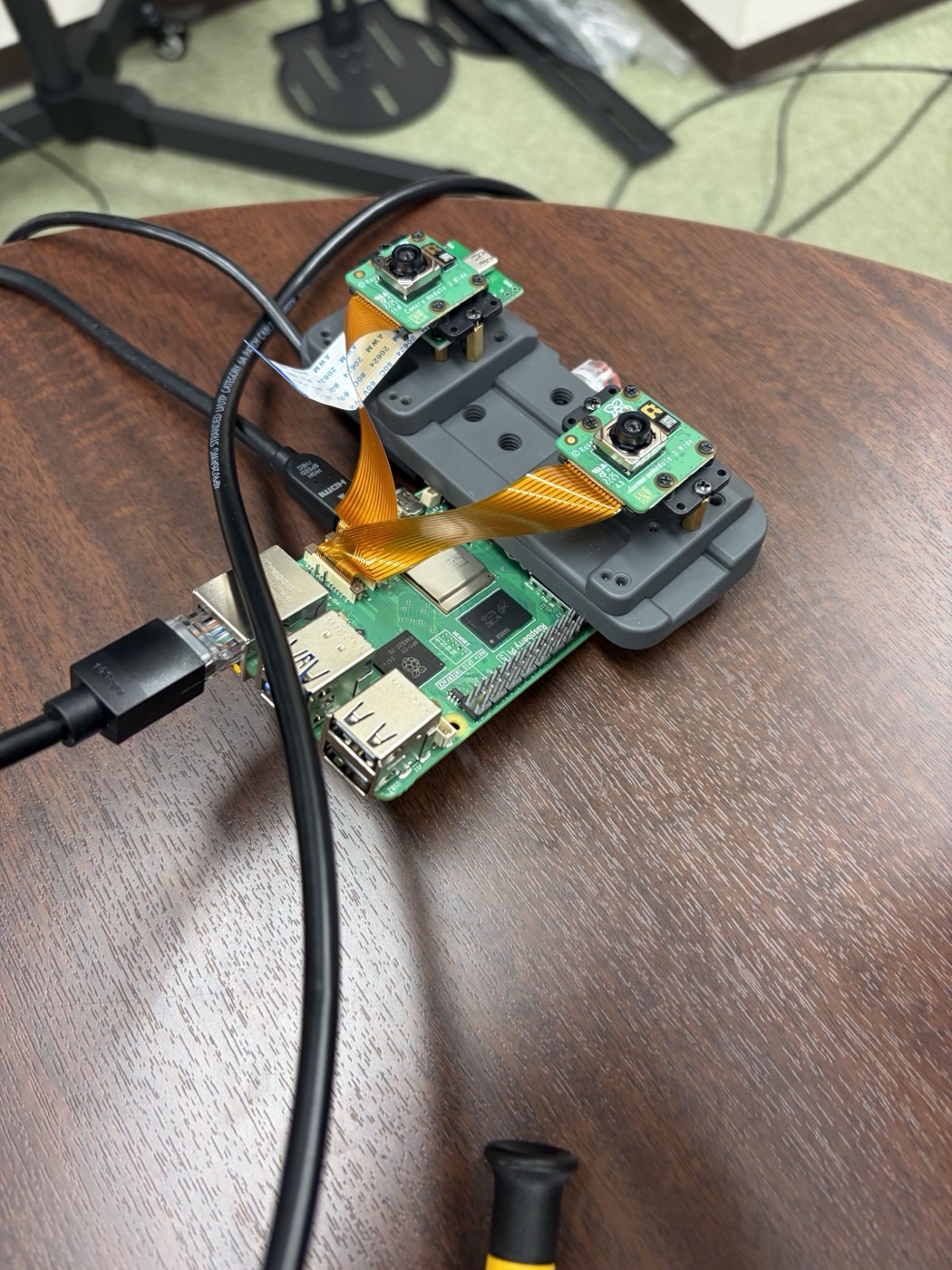 木製の机の上に置かれたRaspberry Pi本体に、2台のカメラモジュールを取り付けたブラケットがオレンジ色のリボンケーブルで接続されている