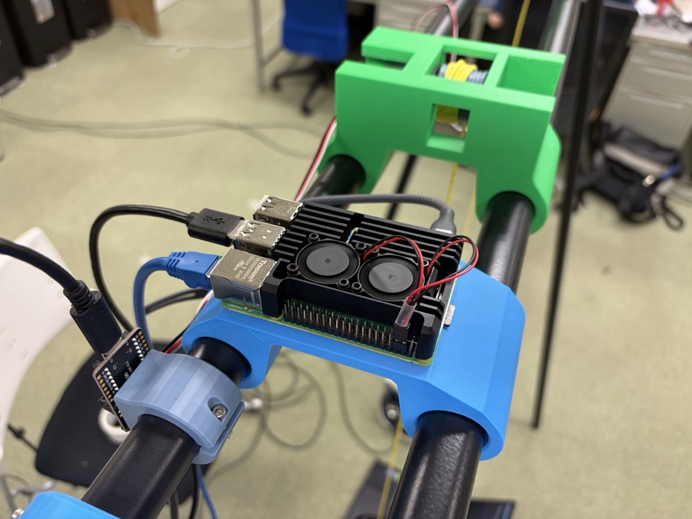 黒いポールに青色のマウントで固定されたRaspberry Pi。ヒートシンクとファンの付いた黒い冷却ケースが載っている。奥には緑色のモーターマウントがクロスバーに取り付けられ、黄色いケーブルが通されている