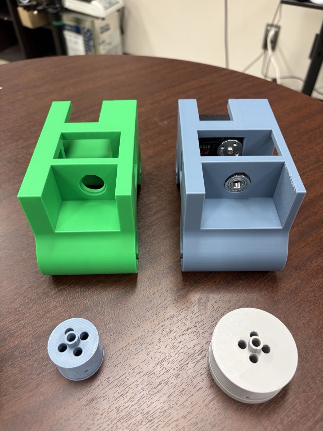 机の上に並んだ2つの3Dプリントマウント。左の緑色のマウントは内部が空洞で軸受けがない。右の灰色のマウントにはサーボが実装され、反対側の軸にボールベアリングが取り付けられている。手前には小さい青色のドラムと、一回り大きい白色のドラムが置かれている